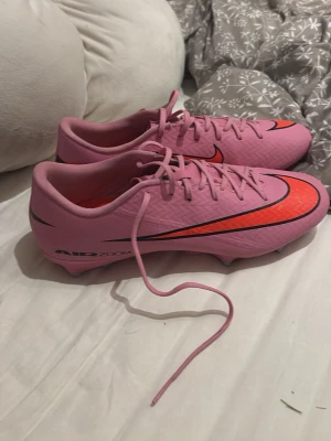 Rosa Nike fotbollsskor Air Zoom - Säljer ett par snygga rosa Nike fotbollsskor med Air Zoom-teknologi. Skorna har en stor svart och röd Nike swoosh på sidan och detaljer i svart och rött. Låg profil och snörning för bra passform. Perfekta för dig som vill sticka ut på planen! OBS‼️‼️‼️ skorna är helt nya endast använda cirka 2 gånger. 