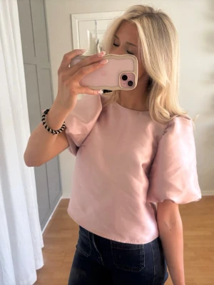 Ljusrosa topp med puffärmar - Söt ljusrosa topp med puffiga kortärmar och rund halsringning. Toppen har en croppad passform och är tillverkad i ett slätt, satinliknande material som ger en elegant känsla. Perfekt för dig som vill ha en trendig och stilren look. Helt ny, aldrig använd, prislapp kvar. 