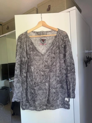 Gråmönstrad blus med v-ringning - Säljer en grå blus med vitt paisleymönster och v-ringad hals. Blusen har långa ärmar och ett avslappnat fall. Materialet känns lätt och luftigt, perfekt för en cool och stilren look. Märket syns på insidan men är otydligt. Från märket Odd Molly i storlek 3 som enligt deras storlekguide motsvarar en storlek L. Mycket fint skick, jag hittar inga synliga anmärkningar eller defekter. 