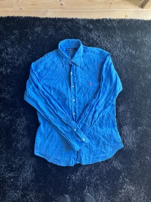 Blå linneskjorta från Ralph Lauren - Säljer en blå långärmad skjorta från Ralph Lauren i linne. Skjortan har klassisk krage, knappar framtill och den ikoniska rosa logotypen broderad på bröstet. Perfekt för dig som gillar en avslappnad men ändå stilren look.