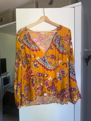 Bohemisk blus i paisleymönster - Säljer en bohemisk blus i härligt senapsgul färg med färgglatt paisleymönster i rött, blått, vitt och grönt. Blusen har v-ringning, långa ärmar med resår vid ärmslut och en lös, luftig passform. Perfekt till både vardag och fest! Använd men i fint skick. Fråga gärna om fler bilder eller mått. Från märket Odd Molly i storlek 3 som enligt deras storlekguide motsvarar en storlek L. Mycket fint skick, jag hittar inga synliga anmärkningar eller defekter. 