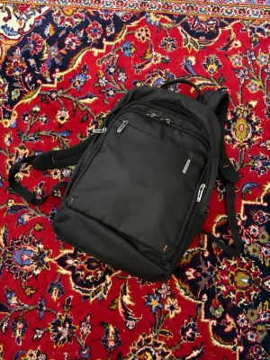 Svart ryggsäck från Samsonite - Snygg svart ryggsäck från Samsonite med flera dragkedjefack och justerbara axelband. Tillverkad i slitstarkt syntetmaterial och har en stilren, modern design med metallogo framtill. Perfekt för skola, jobb eller resor.