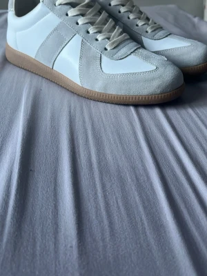 Gats Maison margiellas skor  - Säljer ett par stilrena sneakers i vitt och ljusgrått med snygga mockadetaljer och vita skosnören. Skorna har en klassisk låg siluett och en beige gummisula som ger en clean look. Perfekta för dig som gillar minimalistisk och tidlös stil.