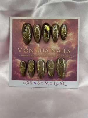 GoldenNight (S):Medium Almond - Unika handgjorda press-on naglar i brun/guldig cateye med guldiga julmotiv som snögubbe, julgran, stjärnor och ornament. Medium Långa, mandelformade naglar som ger en lyxig och kreativ look. Perfekt för dig som vill sticka ut med detaljerade och konstnärliga naglar.S: 15 mm, 11,5 mm, 12 mm, 11 mm, 9 mm 