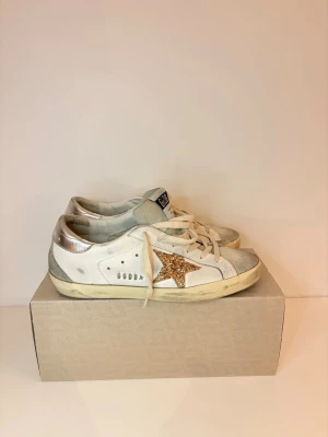 Vita Golden Goose  - Säljer ett par golden goose med en glittrig guldstjärna på sidan och silverdetaljer vid hälen. Skorna har vita snören och en något smutsad sula för en cool, avslappnad look. Perfekta för dig som gillar att sticka ut med detaljer! Dessa skor är asså använda 1, MAX 3 gånger då de inte va min typ av sko, alltså är dem helt helt nya, låda och allt dustbag och sådant tillkommer såklart !! Köp snabbt och smidigt ✨