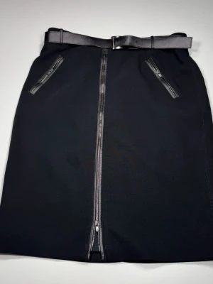 Armani Collezioni Svart kjol med bälte och dragkedja - Stilren svart kjol med markerat bälte i midjan och dragkedja framtill. Kjolen har två fickor med dragkedjor och en rak, klassisk form som passar till många olika looks. Perfekt för dig som gillar minimalistisk och clean stil. 0070