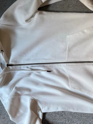 Vit hoodie med dragkedja - Säljer en stilren vit hoodie från JACK & JONES i storlek S. Hoodien har dragkedja framtill, två fickor och justerbar huva med snören. Tillverkad i mjukt bomullsmaterial som känns skönt mot huden. Perfekt för en avslappnad look.