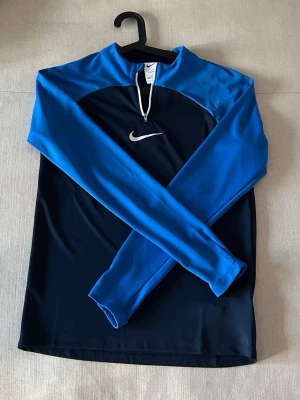 Nike blå långärmad träningströja - Nike långärmad träningströja i blått och mörkblått med vit dragkedja vid halsen och klassisk Nike-logga på bröstet. Tillverkad i snabbtorkande Dri-FIT-material som passar perfekt till träning och sportaktiviteter.