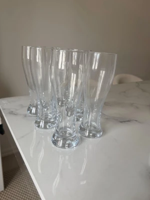 Sex höga ölglas i klart glas - Säljer ett set med sex höga ölglas i klart glas. Glasen har en klassisk, avsmalnande form med bred fot och smalare öppning upptill. Perfekta för att servera öl eller andra drycker. Stilrena och passar bra till hemmabaren eller middagsbordet.