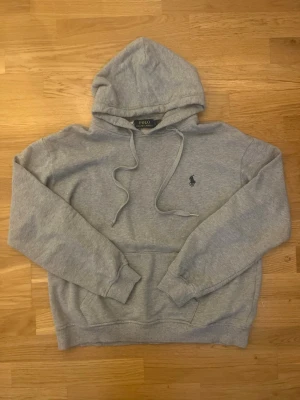 Grå hoodie från Polo Ralph Lauren - Snygg grå hoodie från Polo Ralph Lauren med klassisk huva och dragsnören. Har en broderad logga på bröstet och en stor magficka. Tillverkad i mjukt material som känns skönt mot huden. Perfekt för en avslappnad och stilren look. Storlek L, men sitter defenetivt mer som M. Hör av er vid frågor!
