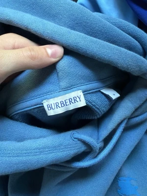Blå hoodie från Burberry - Snygg blå hoodie från Burberry med klassisk huva och dragsko. Tillverkad i mjukt bomullsmaterial som känns skönt mot huden. Perfekt för dig som vill ha en stilren och exklusiv look med diskret logga på bröstet. köpt på NK Collage