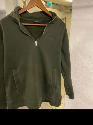 Olivgrön hoodie med halv dragkedja - Snygg olivgrön hoodie från Lindbergh med halv dragkedja framtill och två fickor. Tillverkad i mjukt material och har en klassisk, avslappnad passform. Perfekt för en chill och stilren look. Storlek L