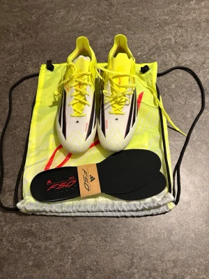 Adidas F50 vita och gula fotbollsskor - Säljer ett par Adidas F50 fotbollsskor i vitt och neon-gult med svarta och röda detaljer. Skorna har snörning, låg profil och levereras med två olika innersulor samt en matchande gympåse. Perfekta för snabba spelare som vill sticka ut på planen.