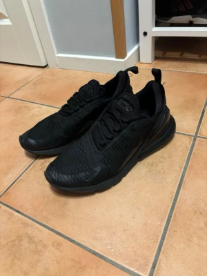 Nike 270 - Säljer ett par helsvarta sneakers med ventilerande mesh-ovandel och kraftig sula. Skorna har snörning, diskreta detaljer och en modern, sportig look. Perfekta för dig som gillar stilrena och bekväma sneakers.