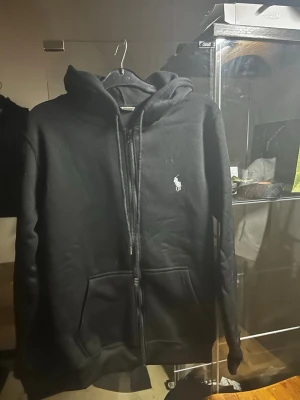  Svart Ralph Lauren, Zip up  - Säljer en svart hoodie med dragkedja från Polo Ralph Lauren. Klassiska design med huva, två fickor framtill och den ikoniska vita loggan broderad på bröstet. Perfekt för en avslappnad och stilren look.