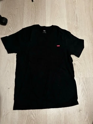 Svart t-shirt från Levi's - Klassisk svart t-shirt från Levi's med liten röd logotyp på bröstet. T-shirten har rund halsringning och korta ärmar. Tillverkad i mjuk bomull för en skön känsla och enkel stil.