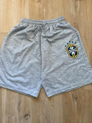 Grå shorts med Brasilien-tryck - Snygga grå shorts med elastisk midja och snörning. På vänster ben finns ett stort Brasilien-inspirerat tryck i gult, blått och grönt. Perfekta för en avslappnad stil eller sportiga dagar. Mjukt material som känns skönt mot huden.