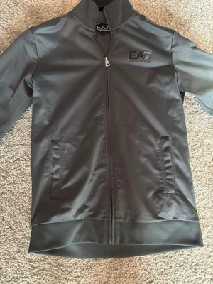 Grå zip tröja från EA7 Emporio Armani - Snygg grå zip tröja från EA7 Emporio Armani med hög krage och hel dragkedja framtill. Den har två fickor och diskret logga på bröstet. Tillverkad i ett slätt syntetmaterial som ger en modern look.