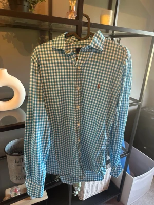 Blåvit rutig skjorta med brodyr - Säljer en långärmad skjorta i blåvitt rutigt mönster med klassisk krage och knappar framtill. Skjortan har en broderad detalj på bröstet och är tillverkad i mjuk bomull. Perfekt för dig som gillar stilrena och tidlösa plagg.