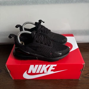 Nike Air Max 270 svarta sneakers - Snygga helsvarta Nike Air Max 270 sneakers med mesh-ovandel och synlig Air-enhet i hälen. Skorna har en sportig siluett, svarta snören och diskreta vita detaljer på sidan och tån. Perfekta för dig som gillar streetwear och vill ha en clean look.