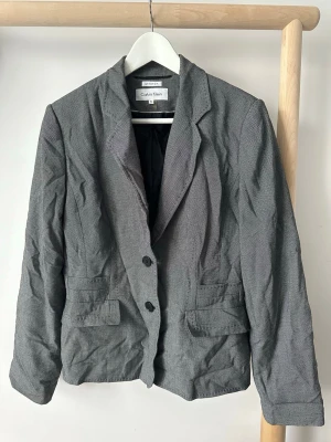 Grå kavaj från Calvin Klein - Snygg grå kavaj från Calvin Klein med två knappar framtill och klassisk krage. Kavajen har två fickor med lock och är tillverkad i ett tunt, strukturerat tyg.  USA storlek 10, motsvarar runt storlek M.