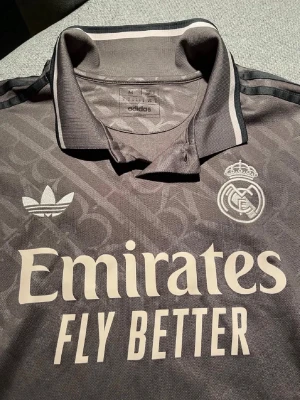 Grå Real Madrid Adidas tröja - Säljer en grå Real Madrid tröja från Adidas med klassisk krage och tryckta loggor. Tröjan har vita detaljer, klubbmärke och Adidas-logga på bröstet samt 'Emirates Fly Better' tryckt framtill. Perfekt för fotbollsfans som vill sticka ut på planen eller läktaren.