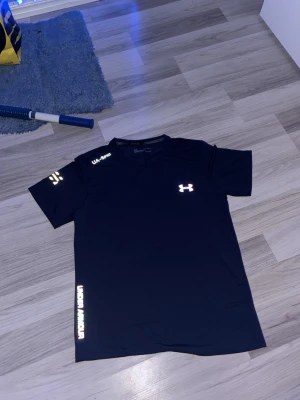 Svart tränings-tshirt Under Armour - Snygg svart tränings-tshirt från Under Armour med reflekterande loggor på bröstet, axeln och längs sidan. Tillverkad i ett lätt och snabbtorkande funktionsmaterial som passar perfekt till gymmet eller löpning.