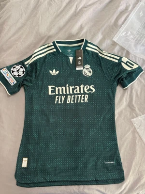  Real Madrid 26/27 Mbappe version - Snygg grön Real Madrid matchtröja från Adidas med vita detaljer och tryck. Tröjan har klubbmärke, Adidas-logga och sponsortryck framtill samt Mbappé och nummer 10 på ryggen. Korta ärmar, rund hals och klassiska ränder på axlarna. Perfekt för fotbollsfans!