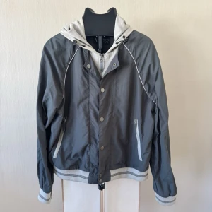 Armani Jeans Grå vindjacka med huva och dragkedja - Snygg grå vindjacka med ljusgrå detaljer och huva. Jackan har dragkedja och tryckknappar framtill, två sidofickor med dragkedja samt ribbade muddar vid ärmslut och nederkant. Perfekt för blåsiga dagar och enkel att matcha till olika outfits.0318 