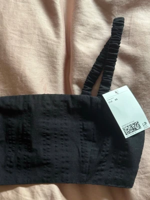 Svart croppad topp - Säljer en svart kort topp med scrunchade axelband. toppen har en enkel, stilren design och är tillverkad i ett mjukt tyg med diskret struktur. Perfekt att bära som en del av ett set. Storlek XS.