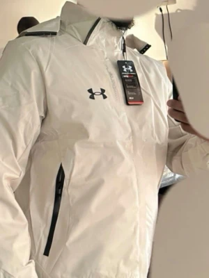 Vit vindjacka från Under Armour - Snygg vit vindjacka från Under Armour med svart logga på bröstet och svarta dragkedjor på fickorna. Jackan har huva och hög krage, samt justerbara detaljer vid huvan. Tillverkad i lätt och vattenavvisande material, perfekt för blåsiga dagar.