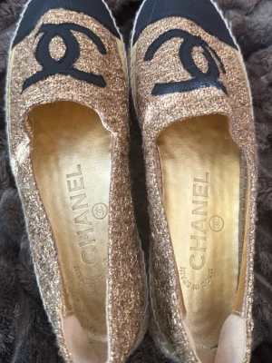 Glittriga guldiga espadrille-sandaler - Säljer ett par espadrille-sandaler i glittrigt guldtyg med svart tåparti och flätad sula i naturmaterial. Skorna har öppen design och är perfekta för dig som vill ha något extra till sommaren. Snygg kontrast mellan guld och svart.