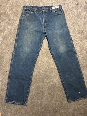 Blå raka Dickies jeans med vintagekänsla - Ett par klassiska blå jeans med rak passform och lätt slitna detaljer. Jeansen har fem fickor och normal midja. Materialet är robust denim i 100% bomull, perfekt för en avslappnad och tidlös stil.