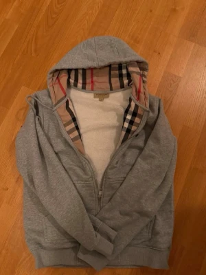 Grå hoodie från Burberry med dragkedja - Snygg grå hoodie från Burberry med dragkedja och klassiskt rutigt foder i huvan. Hoodien har fickor framtill och mjukt, vitt fleecefoder på insidan. Perfekt för dig som gillar exklusiva detaljer och bekväma plagg.