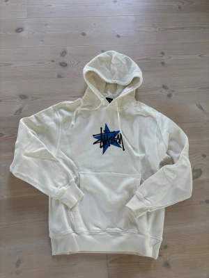 Vit hoodie med blå Stüssy-logo - Säljer en vit hoodie från Stüssy med en stor blå stjärna och svart Stüssy-tryck på bröstet. Hoodien har huva med dragsko, magficka och ribbade muddar vid ärmar och nederkant. Perfekt för en avslappnad streetwear-look.