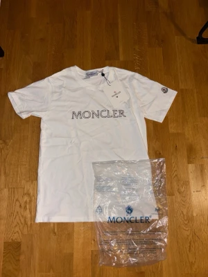 Vit Moncler t-shirt med logga - Säljer en vit t-shirt från Moncler med stor logga tryckt på bröstet och en liten Moncler-patch på ärmen. Klassisk passform och rund halsringning. Perfekt till jeans eller shorts för en clean och stilren look. Storlek S