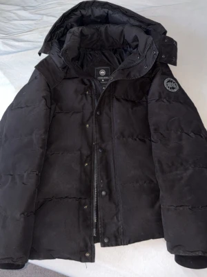 Canada goose black lable - Säljer en svart pufferjacka med avtagbar huva och diskret patch på ärmen. Jackan har dragkedja och knappar framtill samt stora, vadderade paneler för extra värme. Materialet är mjukt och jackan är fodrad med dun och fjädrar för maximal komfort under kalla dagar.