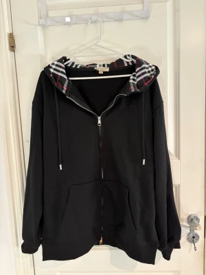 Svart zip hoodie från Burberry - Svart hoodie från Burberry med dragkedja framtill och klassiskt rutigt foder i huvan. Stora fickor på magen och justerbara snören i huvan. Perfekt för en chill och stilren look. Materialet känns mjukt och bekvämt, och detaljerna ger en exklusiv vibe.