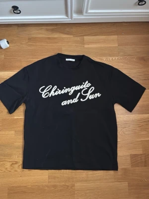 Svart t-shirt med text Chiringuito - Säljer en svart t-shirt med vit text 'Chiringuito and Sun' på bröstet. T-shirten har klassisk passform och korta ärmar. Perfekt för dig som gillar enkel men snygg stil. Tröjan är lite baggy och inte normal passform 