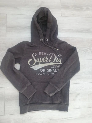 Mörkgrå Superdry hoodie med tryck - Mörkgrå hoodie från Superdry med stort vitt och silverfärgat tryck på bröstet. Klassisk huva med dragsko och magficka framtill. Mjukt material och ribbade muddar vid ärmar och nederkant. Perfekt för en avslappnad och cool stil.