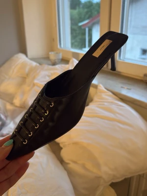 Svarta spetsiga pumps med snörning - Säljer ett par svarta pumps med öppen häl och spetsig tå. Skorna har dekorativ snörning framtill med metallöljetter och en smal klack. Materialet ser ut att vara konstläder med en matt finish. Perfekta för dig som gillar trendiga detaljer.