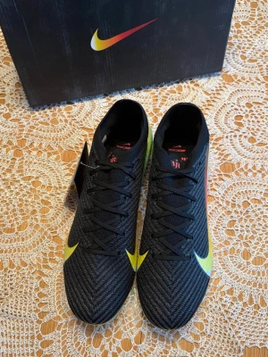 Nike Mercurial fotbollsskor - Säljer ett par Nike Mercurial fotbollsskor i svart med gula detaljer och en gradient swoosh på sidan. Skorna har snörning, låg profil och är tillverkade i ett lätt syntetmaterial för bra bollkontroll. Perfekta för dig som vill ha stilrena och snabba skor på planen.