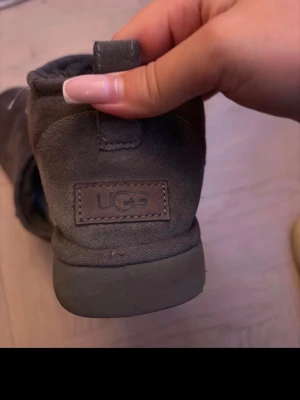 Bruna UGG tofflor i mocka - Mysiga bruna tofflor från UGG i mjuk mocka med låg ankel och tjock sula. Insidan är fodrad för extra komfort och värme. Perfekta för att hålla fötterna varma hemma eller när du vill ha en chill look.