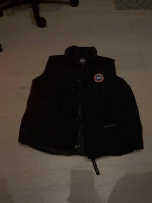 Svart dunväst från Canada Goose - Snygg svart dunväst från Canada Goose med klassisk logotyp på bröstet och hög krage. Västen har dragkedja och tryckknappar framtill samt diskret Canada Goose-brodyr vid fickan. Perfekt för lager-på-lager och håller dig varm under kyliga dagar.