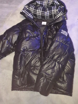 Burberry London England svart dunjacka - Snygg svart dunjacka från Burberry London England. Jackan har en klassisk Burberry-rutig innerfoder i svartvitt och logotyp på ärmen. Perfekt för höst och vinter, håller dig varm och stilren. Storlek L går också att göra till väst 