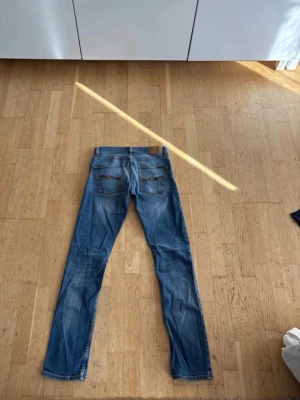 Nudie jeans - Säljer ett par klassiska blå jeans med raka ben och snygga broderade detaljer på bakfickorna. Jeansen har normal midja, bälteshällor och är tillverkade i denim. Perfekta för en avslappnad och trendig stil.