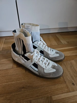 Maison Margiela Performance Sock Gats - Säljer ett par stilrena Maison Margiela sneakers i vitt och svart med klassisk gummisula. Skorna har vita snören, detaljer i mocka och textil samt svarta ränder på sidan. Perfekta för dig som gillar retrovibbar och sportig look.