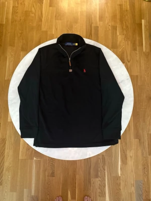 Svart stickad tröja med dragkedja - Svart stickad tröja från Polo Ralph Lauren med halv dragkedja och klassisk röd logga broderad på bröstet. Tröjan har hög krage och ribbade muddar vid ärmslut och nederkant. Perfekt för lager-på-lager och stilren look.