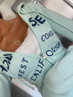 Ljus turkos hollister hoodie med mörkblått broderi - Gullig ljus turkos hollister zipup hoodie med marinblå broderad text över bröstet, strl S omsydd till xxs🪸