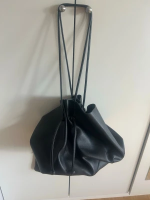 Svart skolväska  - Stilren svart bucket bag i fejkläder med dragskostängning och långa axelband. Väskan har en mjuk, slouchy form och kommer med en matchande liten necessär med dragkedja. Perfekt för dig som gillar minimalistisk och trendig design. Liten väska som passar till skolan💋